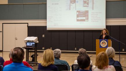 Guest speaker at podium, speaking, with audience in foreground and large screen in background