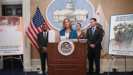 Asm. Irwin speaking, with food containers on podium, and guest speakers in background
