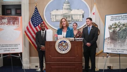 Asm. Irwin speaking, with food containers on podium, and guest speakers in background