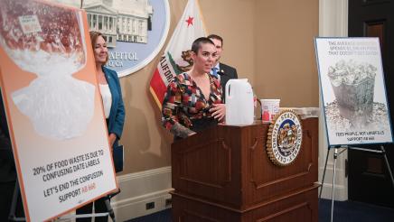 Guest speaker speaking, with food containers on podium as Asm. Irwin and other guest speakers listen on