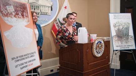 Guest speaker speaking, with food containers on podium as Asm. Irwin and other guest speakers listen on