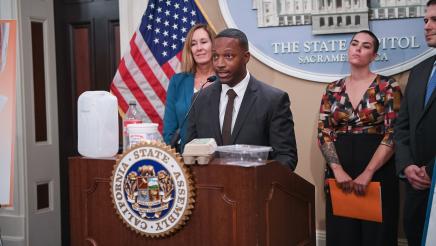 Guest speaker speaking, with food containers on podium as Asm. Irwin and other guest speakers listen on