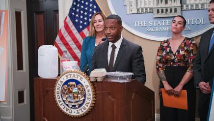 Guest speaker speaking, with food containers on podium as Asm. Irwin and other guest speakers listen on