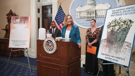 Asm. Irwin at speaking, with food containers on podium and large posters displayed on either side